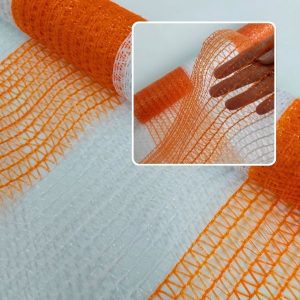 Rolo de tela plástica para tapume nas cores laranja e branco, com detalhe de uma mão esticando a faixa laranja translúcida para evidenciar a malha aberta e a textura do material.