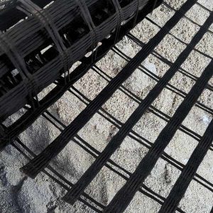 Rolo de geogrelha de fibra de vidro sendo aplicado sobre uma superfície de areia, evidenciando a malha bidirecional com alta resistência à tração