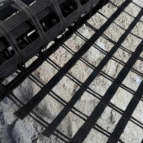Rolo de geogrelha de fibra de vidro sendo aplicado sobre uma superfície de areia, evidenciando a malha bidirecional com alta resistência à tração