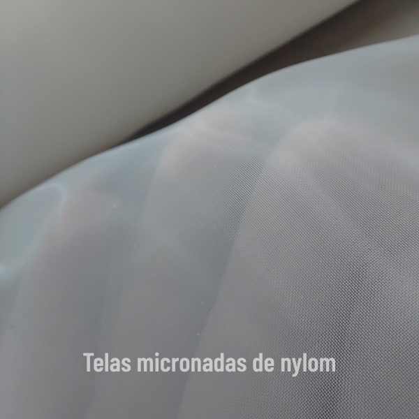 Close de uma tela micronada de nylon branca e translúcida, esticada sobre rolos, com a frase ‘Telas micronadas de nylon’ escrita na parte inferior